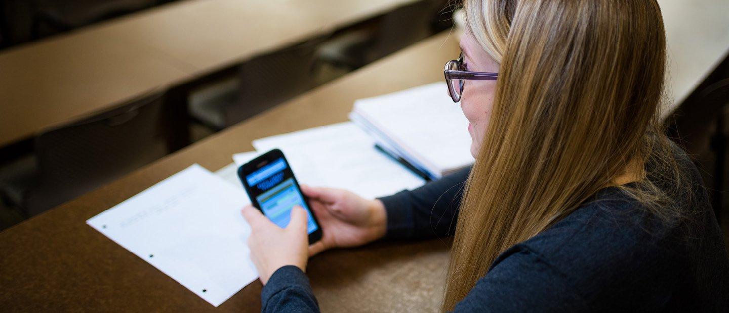 A young woman seated at a table with papers, using a smart phone.